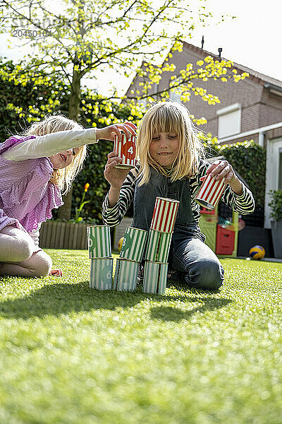 Two children are playing with colorful blocks on a grassy lawn  with one child in the act of placing a block on a tower while the other observes.