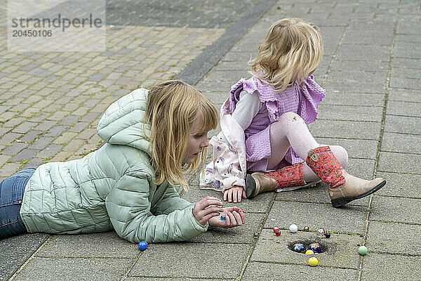 Two children are playing a game with marbles on a paved surface outdoors. One child is in a purple dress sitting while the other is wearing a green jacket and is partially lying down.