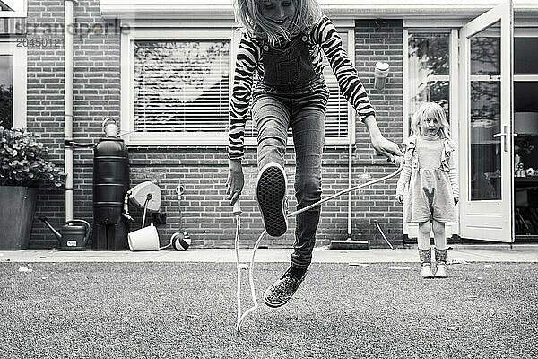 A black and white photo capturing a moment of play  with a girl in mid-air turning a skipping rope and another child watching in the background  set against a residential backdrop.