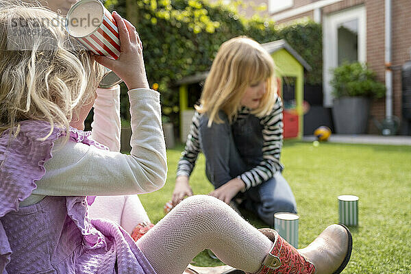 Two young children are playing in a sunlit garden  one drinking from a striped cup and the other in overalls observing.