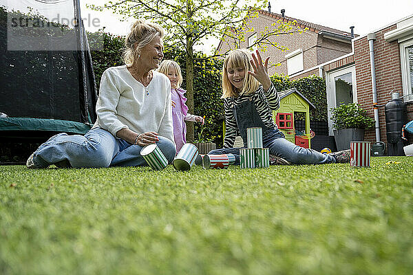 An elderly woman and two young girls are sitting on a grass lawn playing with colorful tin cans  set up like bowling pins  with a child's playhouse in the background.