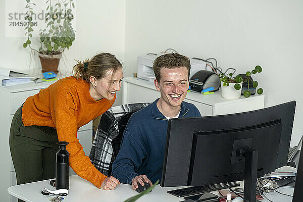 Two individuals are laughing and looking at a computer screen in a bright office setting. One sits at the desk  while the other stands beside them.