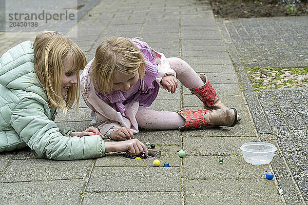 Two children are engrossed in playing with marbles on a paved sidewalk  with a clear plastic container beside them.