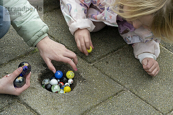 A child and an adult play with colorful marbles on a paved surface  focusing intently on the game at hand.