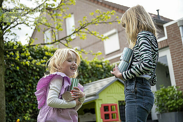 Two children are playing outdoors  one holding a toy and tin cans while laughing with the other who stands with hands on hips.