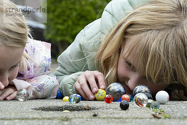 Two children are engrossed in playing with a collection of colorful marbles on the ground.