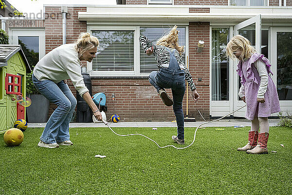 An adult woman is playing with two young girls in a backyard garden  assisting one with a skipping rope while the other watches.