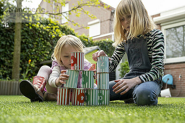 Two young children are focused on stacking multicolored tin cans in a grassy backyard  creating a tower.