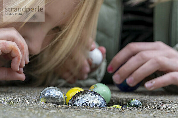 A child is playing with colorful glass marbles on the ground  closely examining one while laying on their stomach.