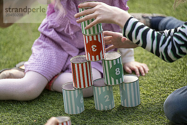 Children playing with a colorful stack of cans designed with numbers and stripes on a grass surface.