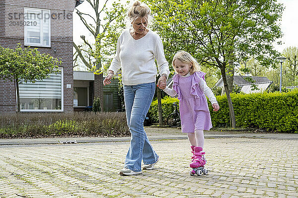 An older woman is assisting a young girl who is learning to skate on rollerblades in a suburban street.