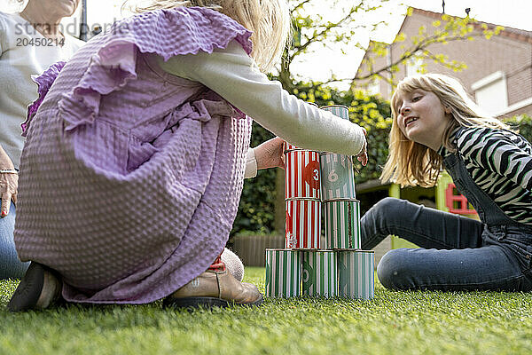 A child in purple clothing and a female teenager in a striped sweater play with colorful tin cans on a grass lawn  under the watchful eye of an adult  whose partial figure is visible.