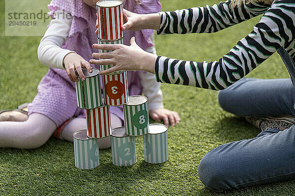 Two children are playing with colorful numbered cans on a grassy surface  stacking them up in a playful outdoor activity.