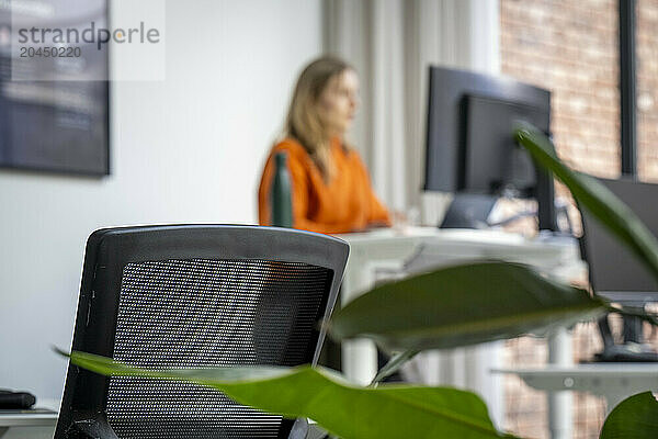 Office environment with a focus on an office chair in the foreground and a woman working at her computer in the background  slightly out of focus.