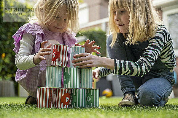 Two young children are engrossed in play outdoors  stacking colorful toy cans in a make-believe game on a lush green lawn.