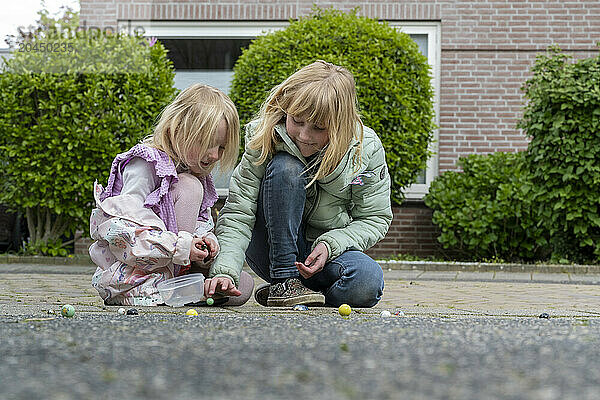 Two young girls are playing with marbles on a sidewalk in front of a brick house.