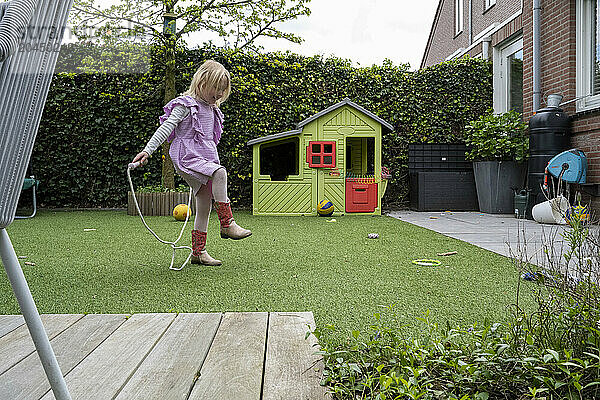 A young child plays in a backyard  dragging a plastic chair across artificial grass near a colorful playhouse.