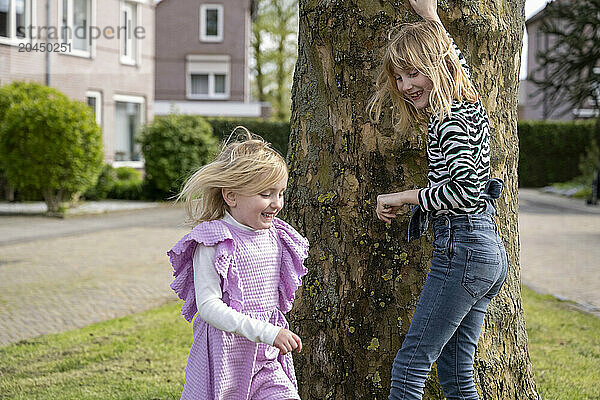 Two children are playing around a tree in a residential area  one hanging from the tree while the other stands beside it  both smiling joyfully.