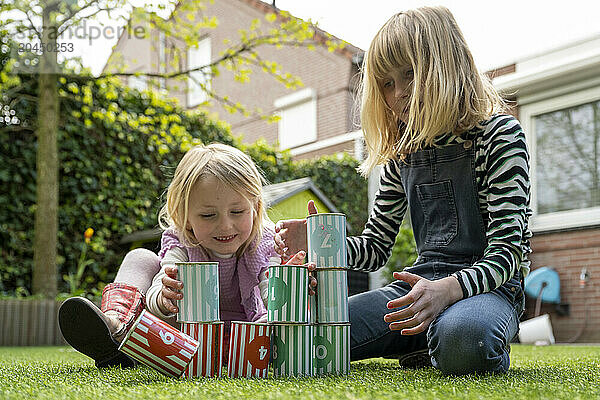 Two children are playing outdoors  stacking colorful tin cans on a lawn with a residential backdrop.