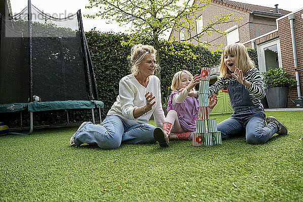 A woman and two children are playing with a tower of colorful blocks in a backyard garden  with expressions of excitement as the blocks fall.