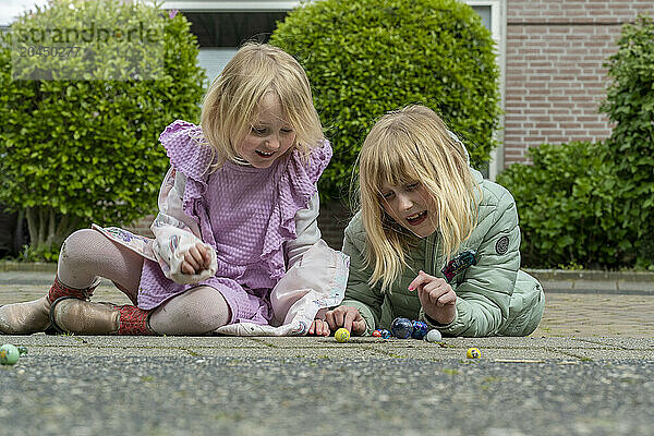 Two children are playing with marbles on a paved surface  appearing delighted and engrossed in their game.