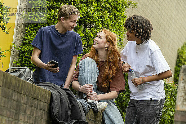 Three teenagers are socializing outdoors  with one showing something on a mobile phone to a smiling girl while another stands by with a beverage in hand.