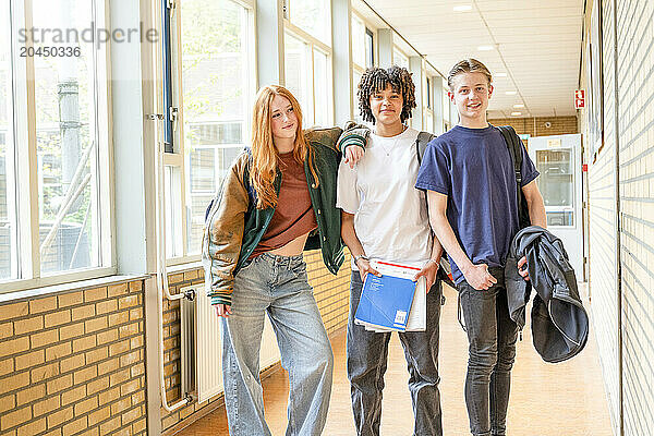 Three teenagers stand in a bright school hallway  smiling and looking directly at the camera. The girl on the left has red hair and the two boys have brown hair  with the middle one having curly hair. They are casually dressed  suggesting a relaxed school environment.