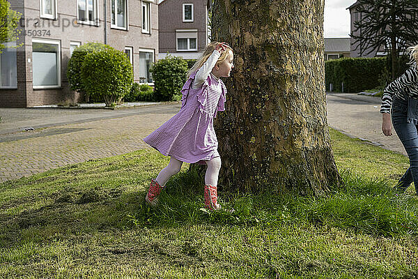 A young girl in a pink dress and red boots is playing hide and seek around a tree on a residential street with grass patches  while an adult woman stands nearby.