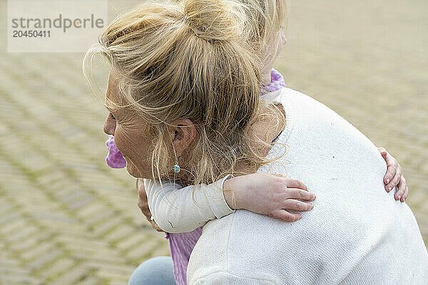 A mother carries her young daughter on her back  both with blonde hair  embracing a tender moment together outdoors.