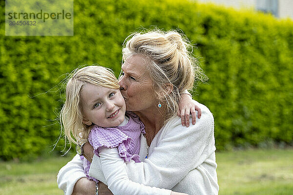 A woman is affectionately kissing a young girl on the cheek outdoors  with a green hedge in the background.