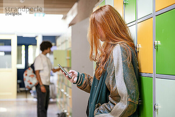 A red-haired teenager is focused on her smartphone in a school hallway with lockers and another student in the background.