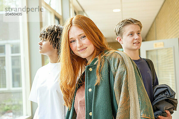 A smiling young redhead woman in casual attire with two male teenagers in the background stand in a school corridor.