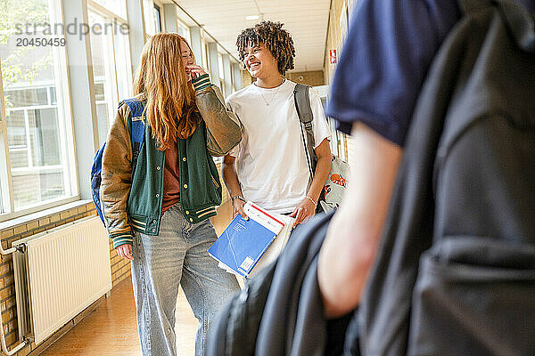 Two teenagers walking down a school corridor  engaged in a cheerful conversation with books in hand.