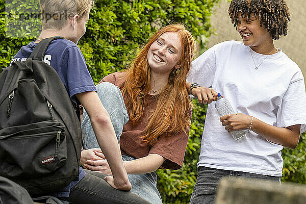 Three teenagers are socializing outdoors  laughing and engaging in a conversation  with one holding a water bottle.