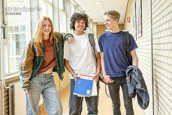 Three smiling students with backpacks stand in a well-lit school corridor holding books and folders.