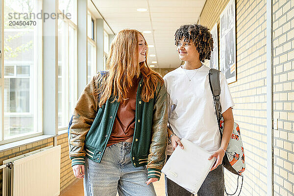 Two students are walking through a school hallway  smiling and conversing with each other. One is carrying a notebook and the other has a backpack over one shoulder.