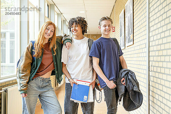 Three students are smiling and posing in a school hallway  each carrying school supplies.