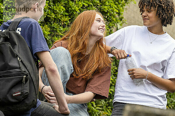 Three friends are engaged in a lively conversation outdoors. A redhead woman is laughing with a woman holding a water bottle  while a man with a backpack looks on.