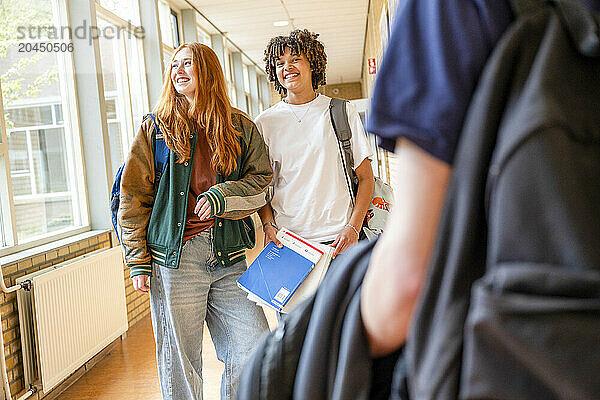 Two students are walking down a school hallway  smiling and engaging in conversation  with books in hand.