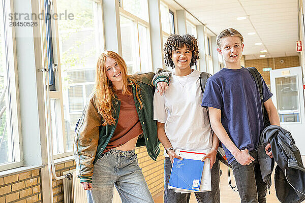 Three students are smiling in a school hallway  each holding various items like books and a backpack.