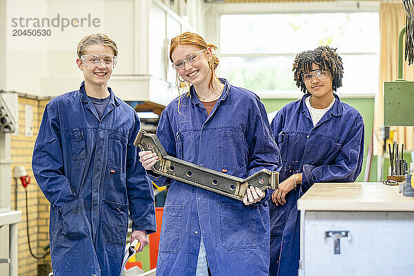 Three apprentices stand side by side dressed in blue work overalls in a workshop  smiling as they hold a large metallic tool together.