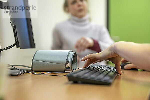 A person's hand is typing on a keyboard with a view of a blurred colleague in the background.