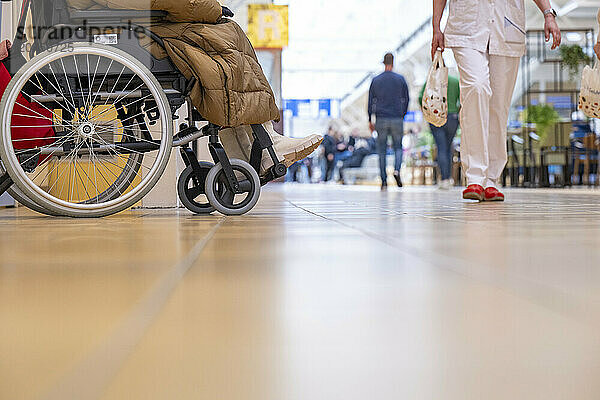 A wheelchair with a beige coat hanging from it is in focus within an indoor facility  while an individual walks away in the background.