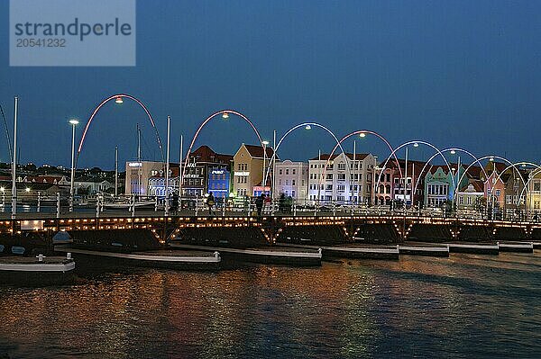 View at dusk of Queen Emma Bridge Queen Emma Bridge swing bridge Emma-Brug floating bridge with 16 pontoon boats Pontjesbrug pontoon bridge movable floating bridge over St Anna Bay Sint Annabaai in Willemstad behind it landmark illuminated trading arcade houses in colonial style in Dutch Dutch Caribbean  Caribbean  Windward Islands  Netherlands Antilles  ABC Islands ABC Islands  Willemstad  Curaçao Curacao