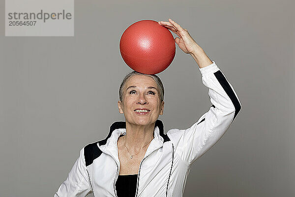 Happy active senior woman balancing red ball on head against gray background