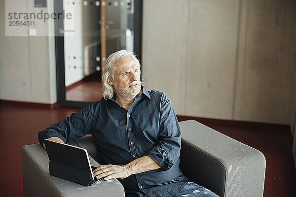 Thoughtful senior businessman with tablet PC sitting on armchair in office