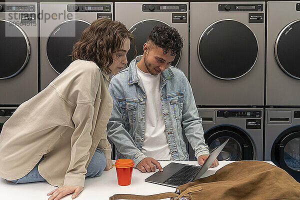 Young couple using laptop on table in front of washing machines at laundromat