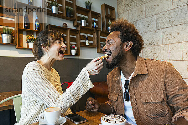 Cheerful woman feeding boyfriend sitting in cafe