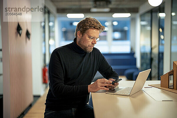 Mature businessman using smart phone and sitting with laptop at desk in office