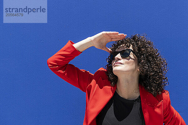 Curly haired woman wearing sunglasses shielding eyes in front of blue wall on sunny day